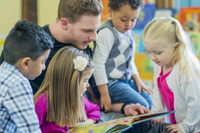 A multi-ethnic group of young children are reading at a preschool with their male teacher.