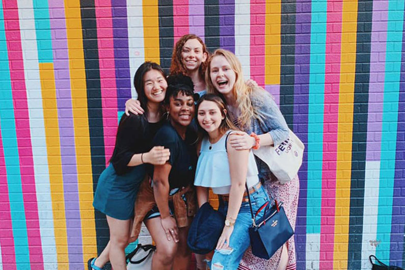 Smiling female students gather in front of vertical colored stripes.