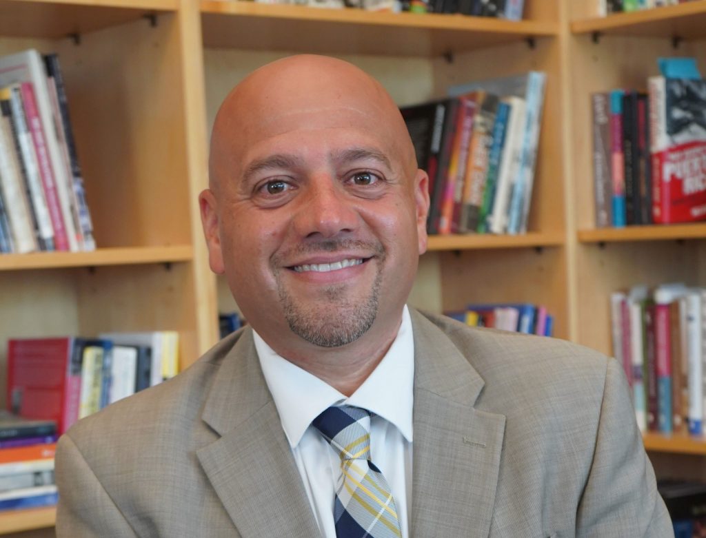 Dean Jason Irizarry sits in front of his office bookcase.