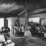 African American children sit in one-room school house around heater in a historic photo.