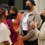 Two women wearing masks stand talking with each other among a crowd.