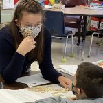 Female teacher wearing mask helps young student.