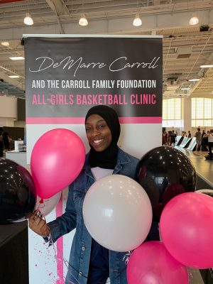 Olusekun stands in front of an all-girls basketball clinic banner with pink and white balloons.