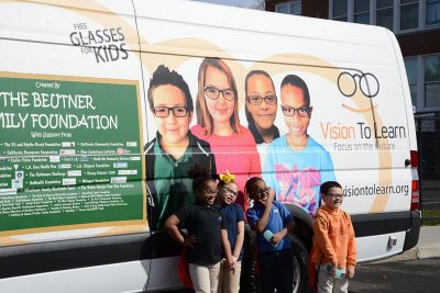 Four young students wearing new eyeglasses stand in front of mobile vision clinic.